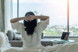 Mujer sentada sobre su cama, estirándose y mirando el paisaje a través de su ventana. Disfrutando de una gran calidad de vida.