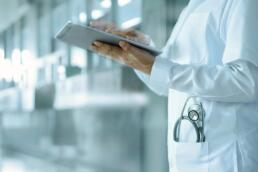 Cuerpo de mujer con bata de doctor, estetoscopio y tableta electrónica, parada en el pasillo de un hospital.