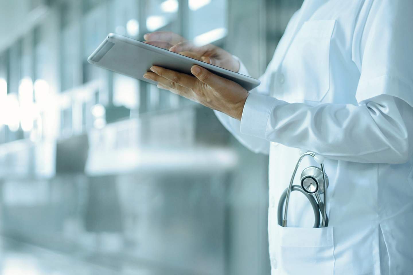 Cuerpo de mujer con bata de doctor, estetoscopio y tableta electrónica, parada en el pasillo de un hospital.