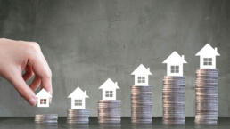 Mano de una persona colocando una casa sobre una torre de monedas ilustrando el aumento del rendimiento al invertir en inmuebles