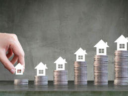 Mano de una persona colocando una casa sobre una torre de monedas ilustrando el aumento del rendimiento al invertir en inmuebles