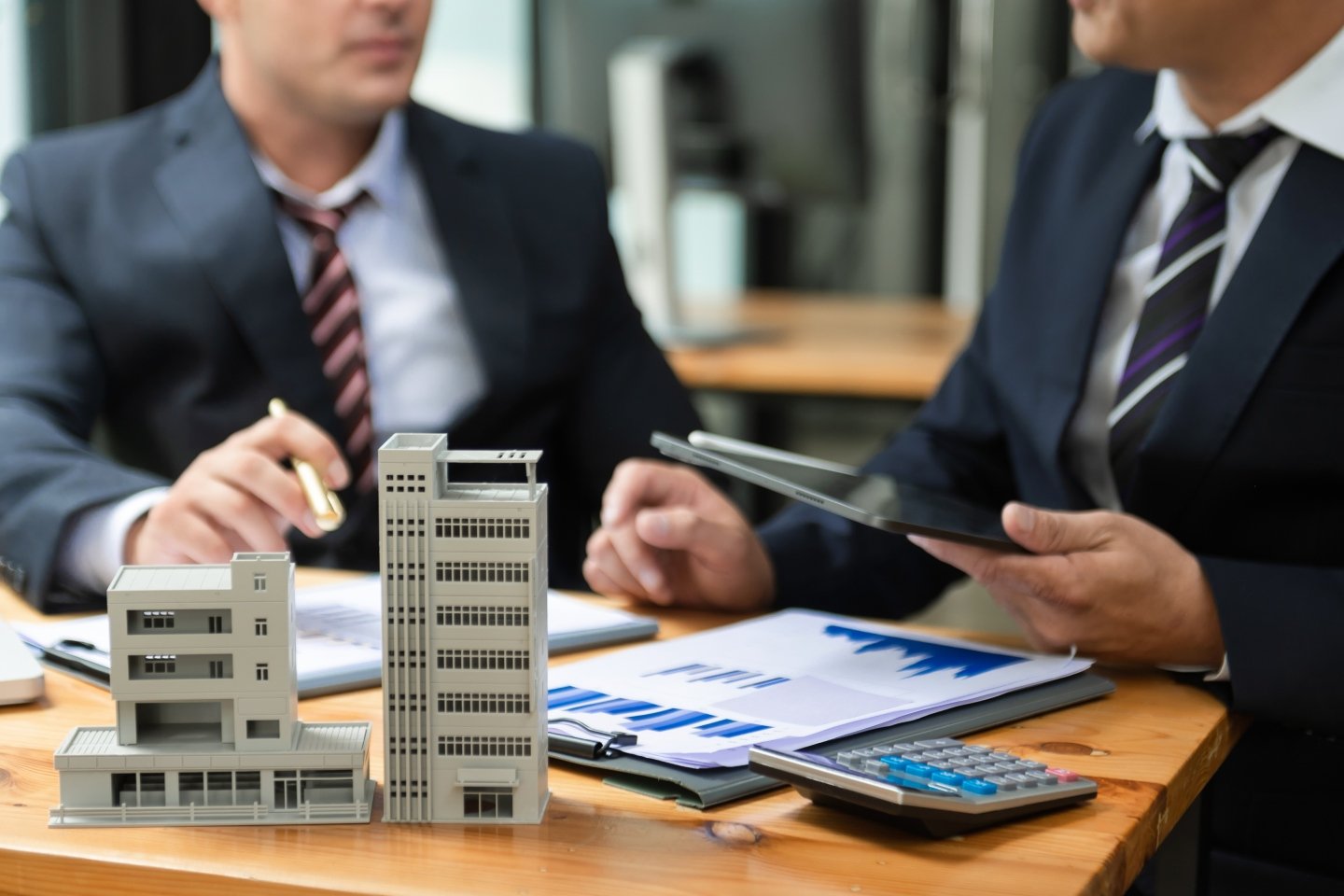 Maquetas de edificios de departamentos sobre una mesa. Con una calculadora, papeles con gráficas y manos de dos hombres al fondo.