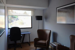 Cuarto de un departamento ambientado como una oficina en casa. Escitorio con silla en frente, lampara a un costado, dos sillones, mesa lateral pequeña con un libro